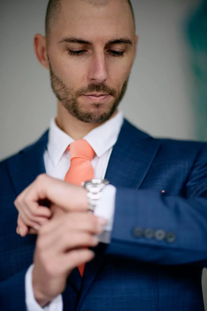 James checking his watch, blue suit, coral tie.