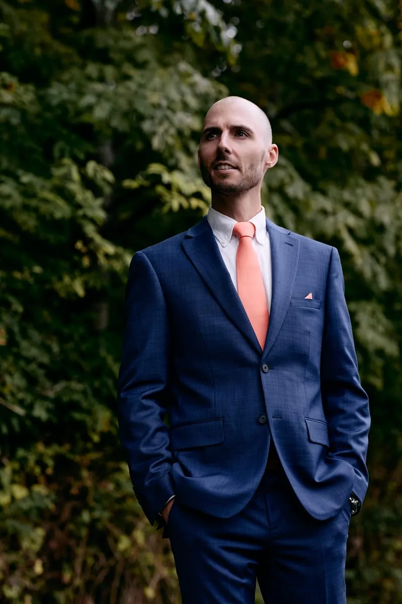 James in the garden, hands in pockets, blue suit against green leaves.