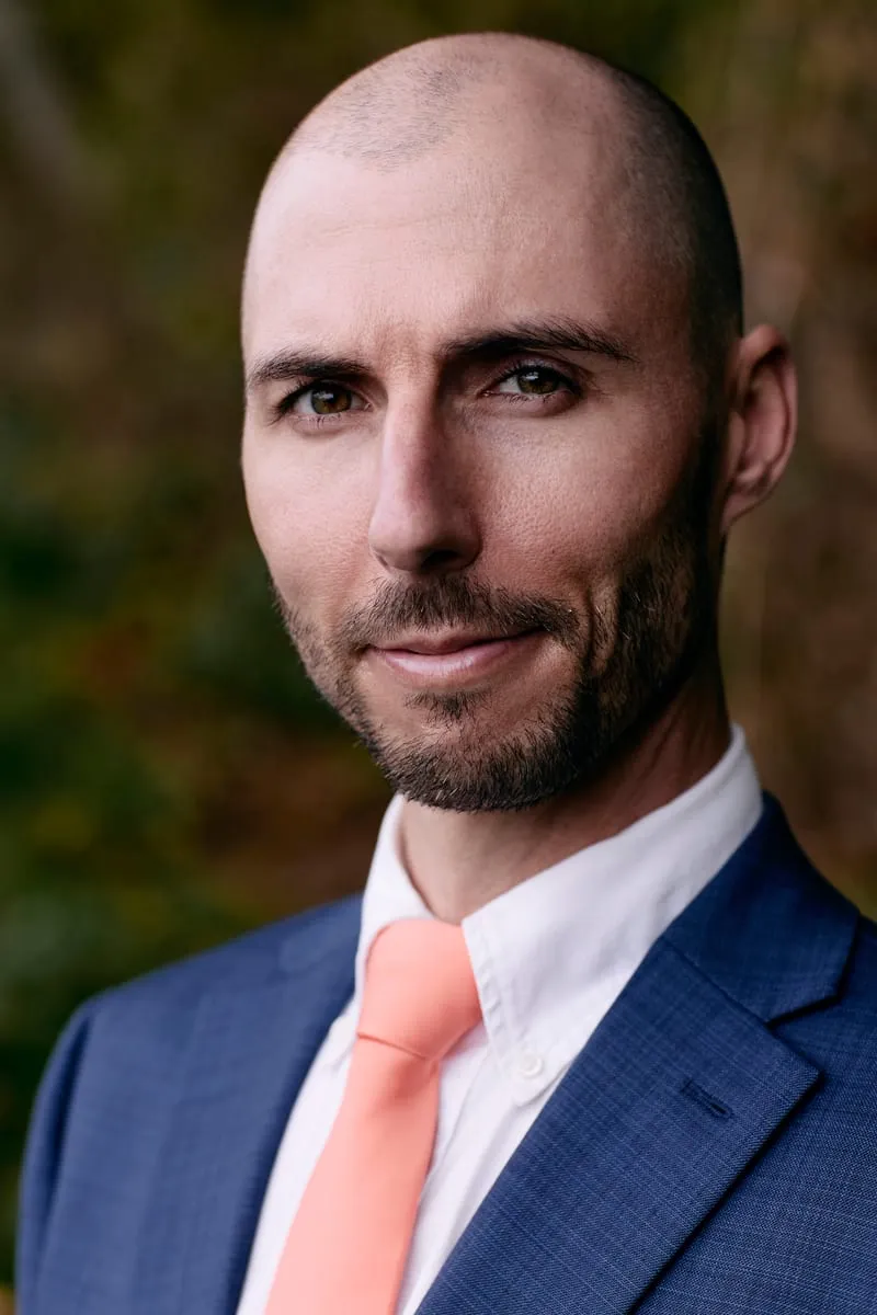 Portrait of James in a blue suit and coral tie against dark leaves.