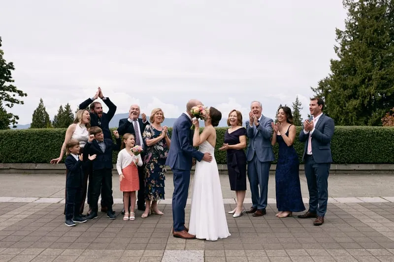 Olivia and James kissing behind her bouquet while their families clap and cheer.