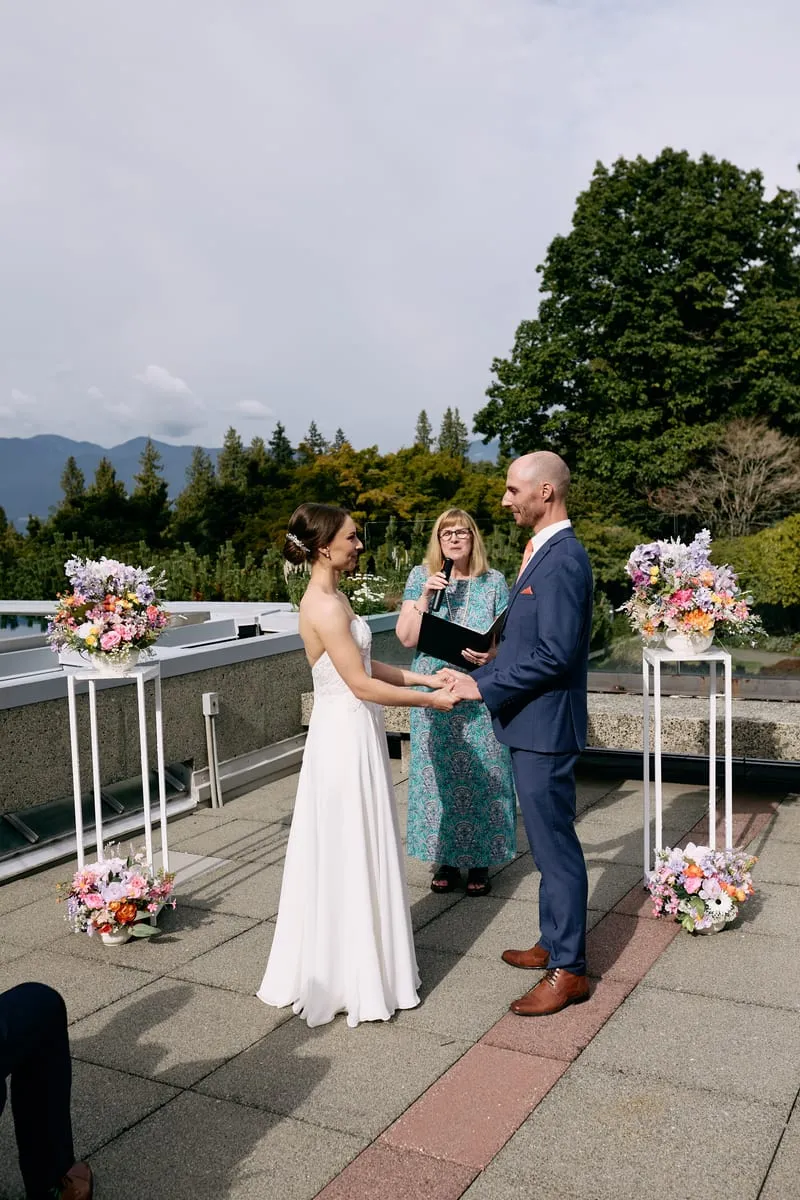 Olivia and James hold both hands between two stands of flowers, the officiant behind them.