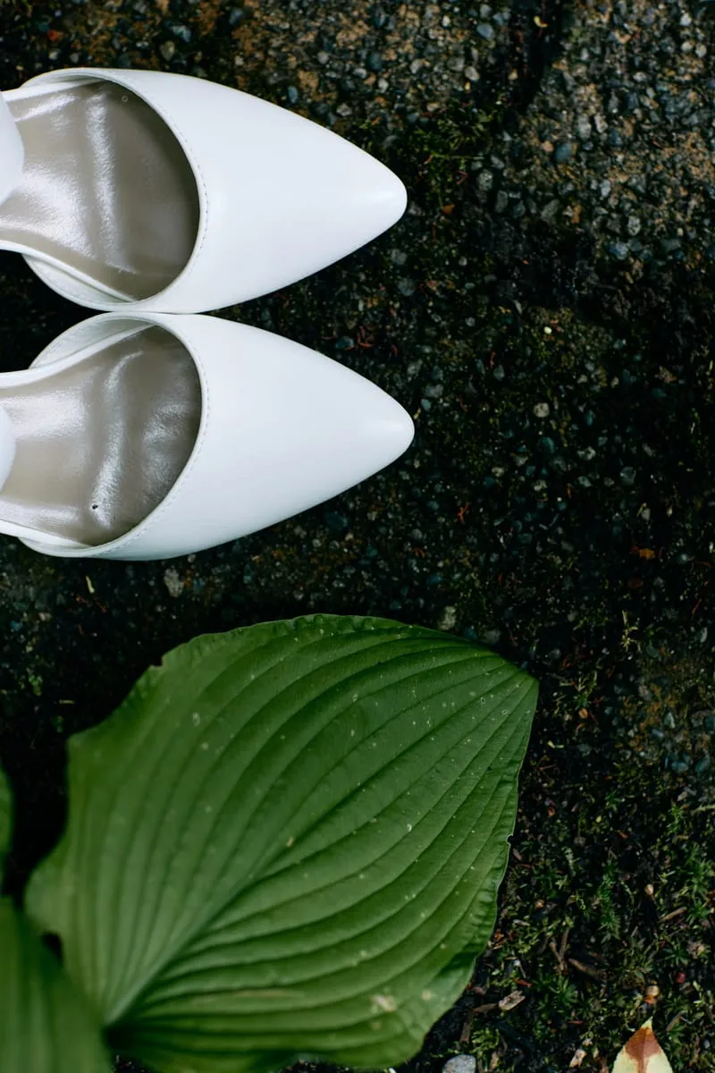 White pointed bridal shoes on dark wet stone beside a broad hosta leaf.