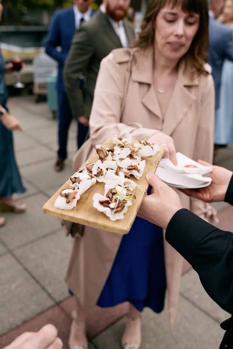 A guest in a trench coat takes a canapé from a wooden board passed around the terrace.