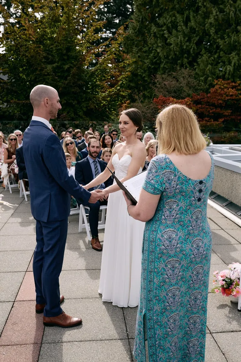 Olivia and James hold hands facing each other while the officiant reads, guests seated behind.