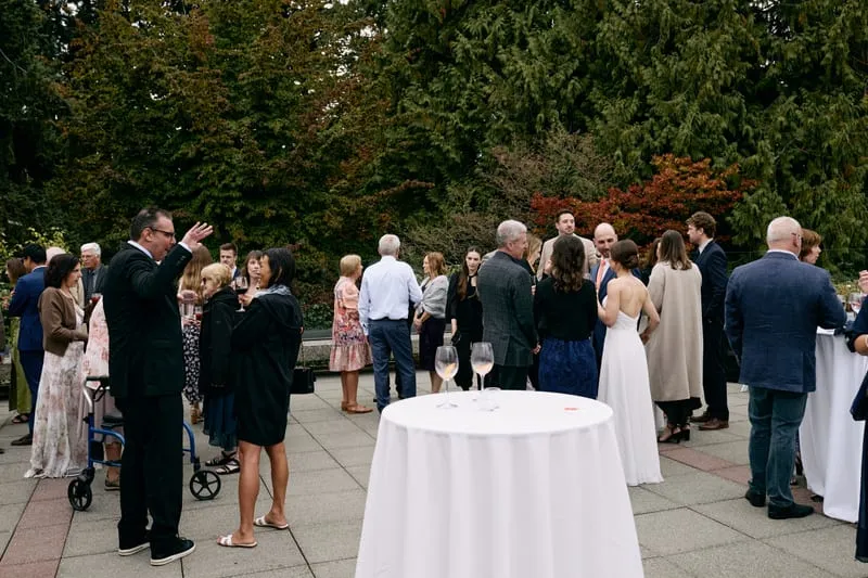 Guests talk around tall white tables on the terrace, trees turning behind them.