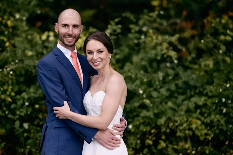 Olivia and James hold each other in front of a dense green hedge, smiling at the camera.
