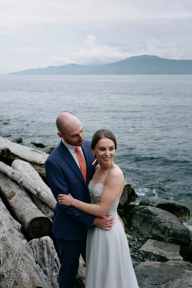 Olivia laughs in James's arms on the driftwood, the water and a far shoreline behind them.