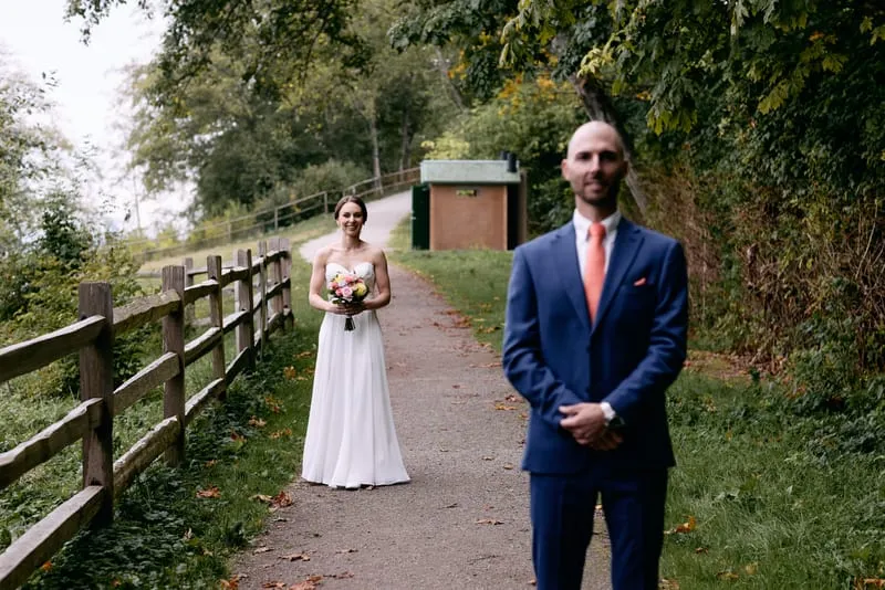 James waits on a fenced forest path with his back turned as Olivia walks up behind him holding her bouquet.