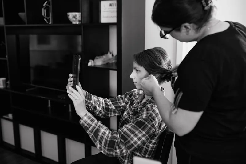 In black and white, a stylist pins up a woman's hair while she checks it on her phone.