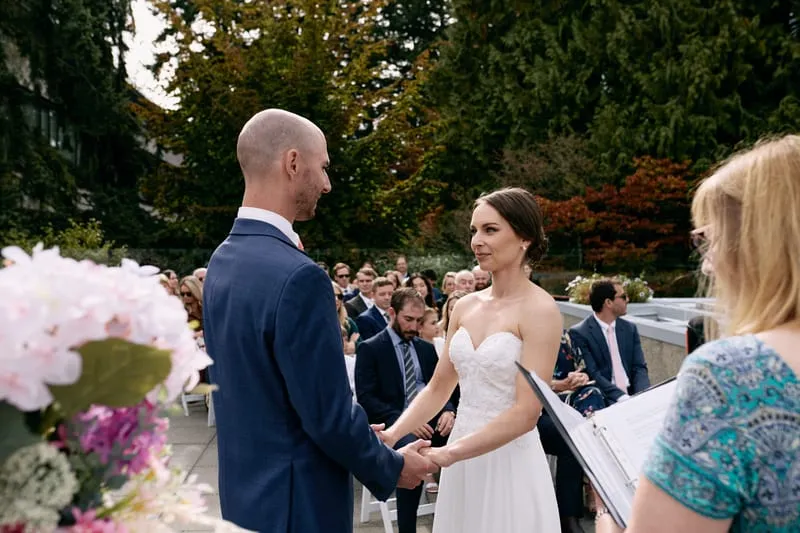 Olivia and James hold hands as the officiant reads from her folder, flowers in the foreground.