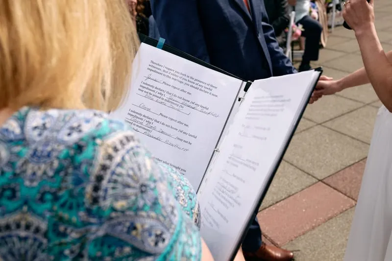 Over the officiant's shoulder, the ceremony script open in a black folder.