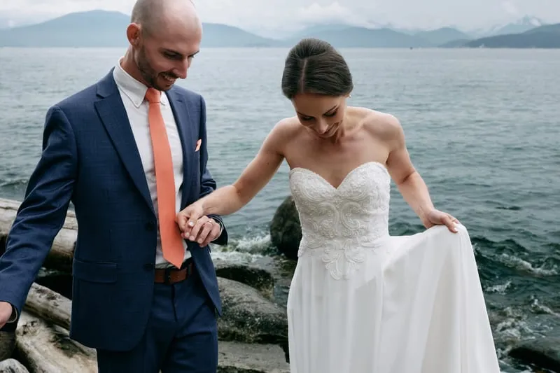 James helps Olivia over the rocks as she lifts her dress, mountains across the water.