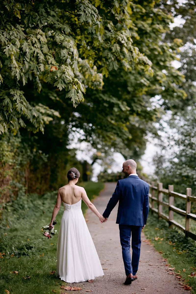Seen from behind, Olivia and James walk hand in hand along a fenced path under tall trees.