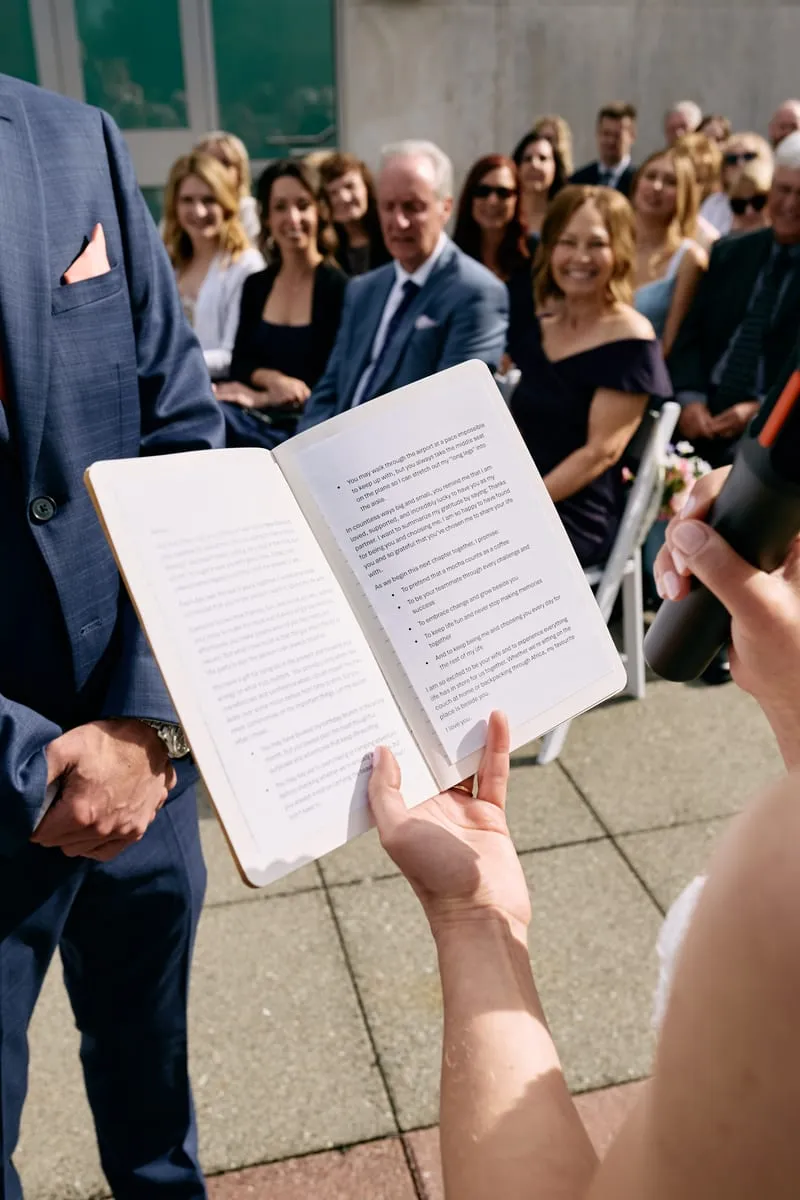Over Olivia's shoulder, her vows in an open notebook, the guests smiling in the rows ahead.