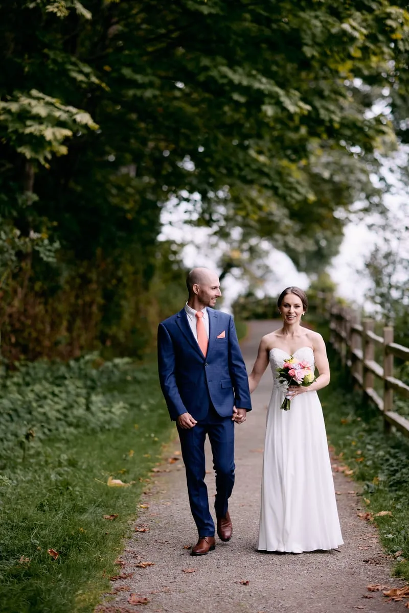 Olivia and James walk hand in hand down a gravel path under the trees.