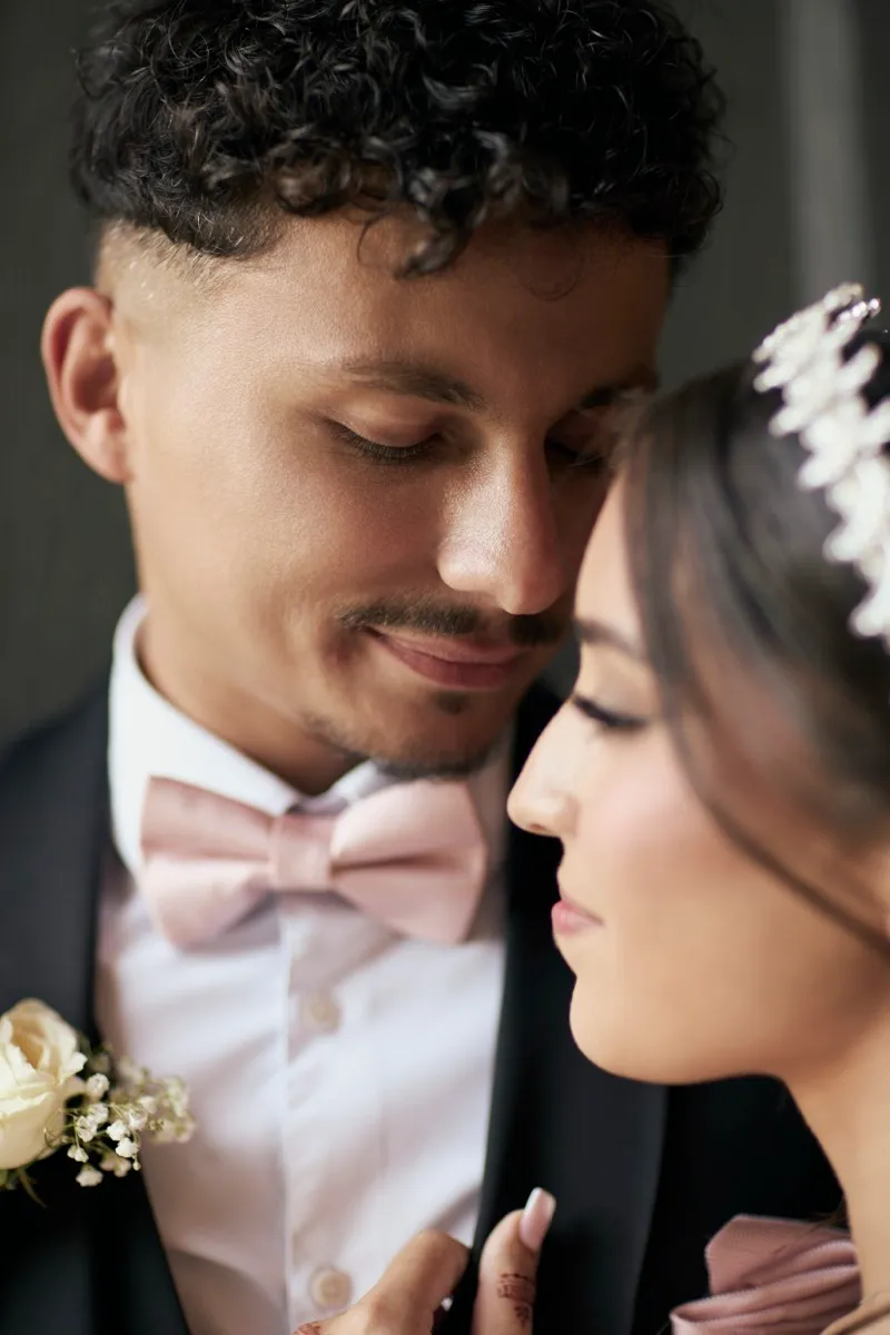 Close portrait of a groom in a pink bow tie with his eyes closed, forehead to forehead with his bride.
