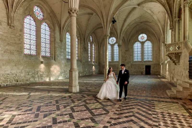 A bride and groom walk hand in hand across the tiled floor of a vaulted Gothic hall with tall stained-glass windows.