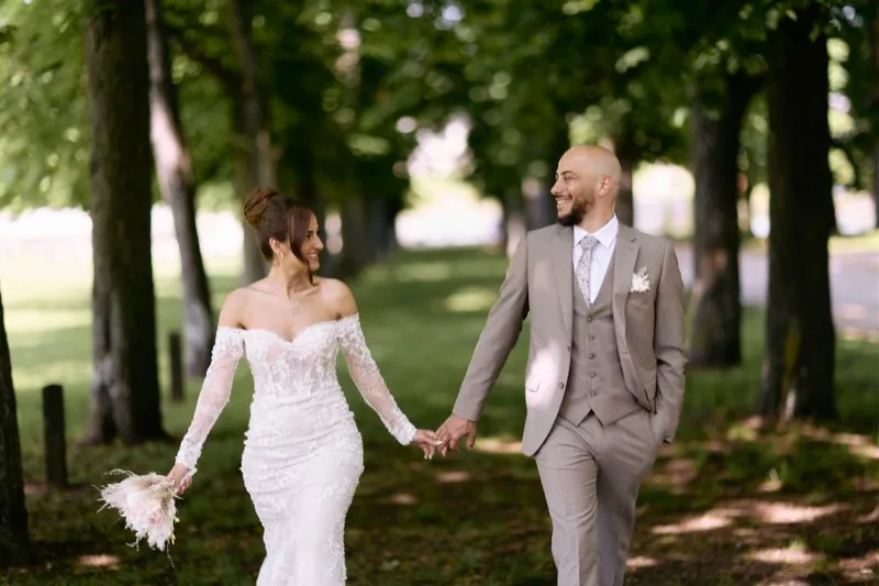 A bride in a long-sleeved lace gown and a groom in a three-piece grey suit walk hand in hand between rows of trees.