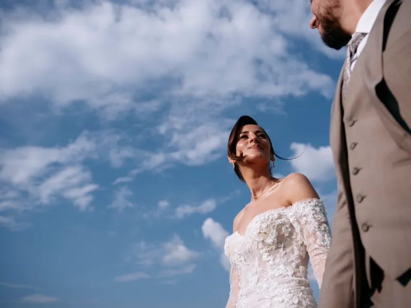 A bride looks up at her groom against a deep blue sky with white clouds, photographed from below.