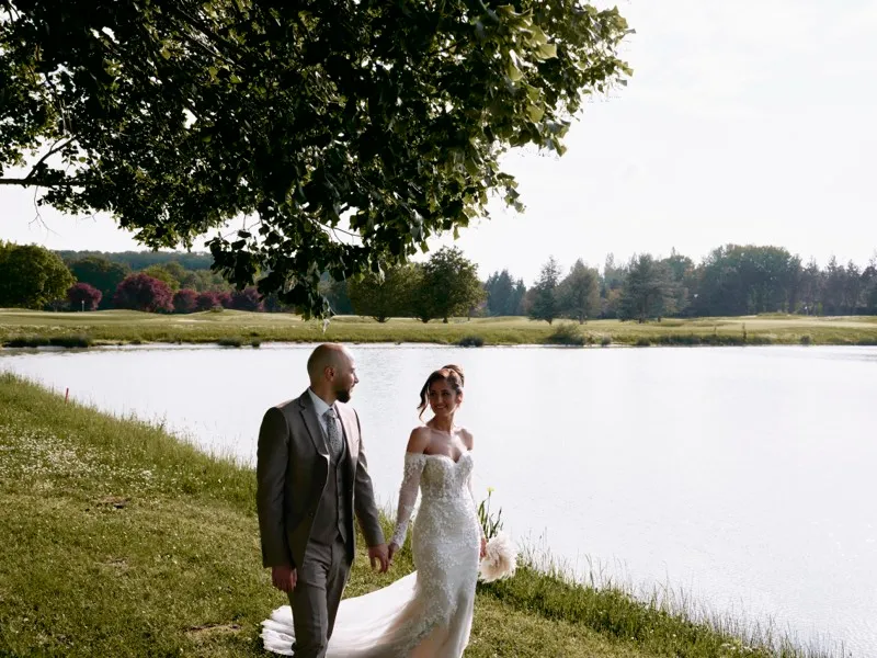 A bride and groom walk hand in hand along the grassy edge of a lake beneath a large tree.