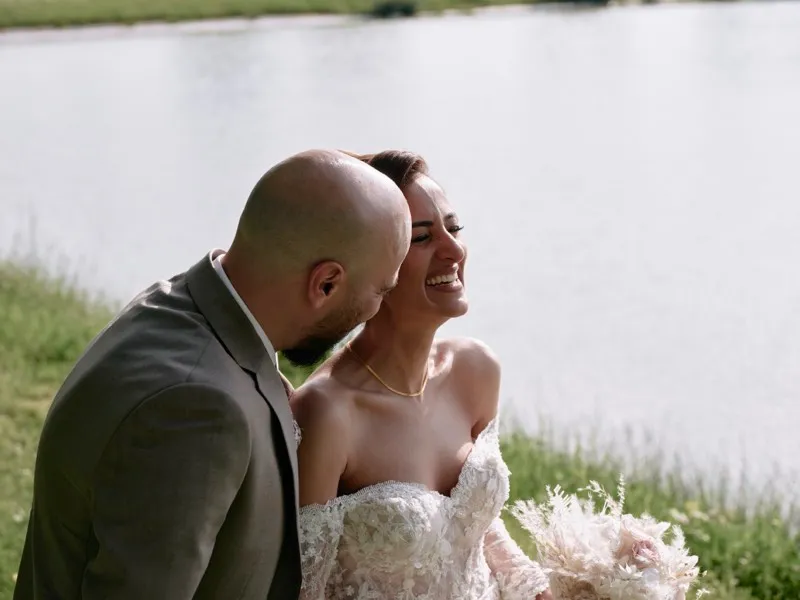A groom kisses his laughing bride's cheek on the grass by a lake, her bouquet in hand.