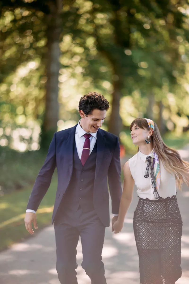 A couple walk hand in hand under an avenue of trees in dappled light, she in a white blouse and patterned skirt.