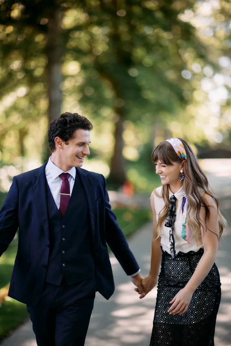 A man in a navy three-piece suit and a woman with a printed headscarf swing their joined hands as they walk under the trees.