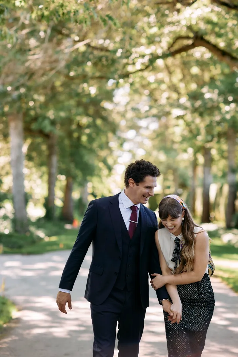 A couple walk arm in arm and laughing down a path beneath an arch of trees.