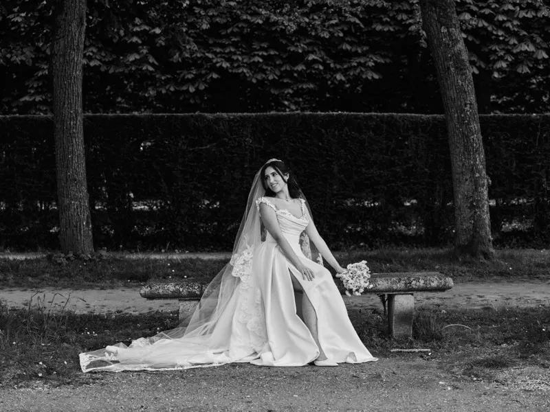 Black and white photograph of a bride in a full gown and veil seated on a stone bench in front of a tall clipped hedge.