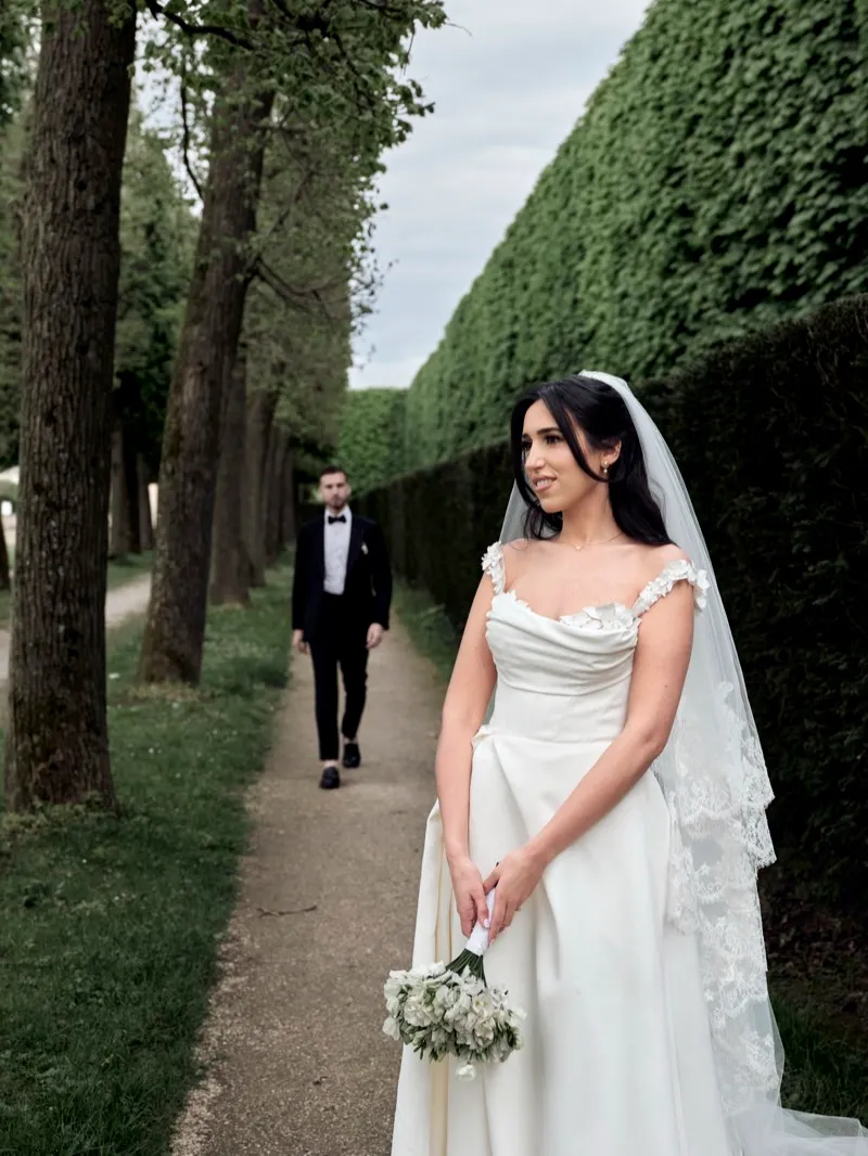 A bride in a satin gown and lace veil walks toward the camera down a path between tall hedges while the groom follows behind.