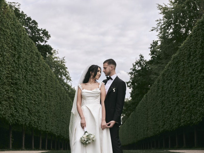 Bride and groom walking a tree-lined avenue in formal gardens.