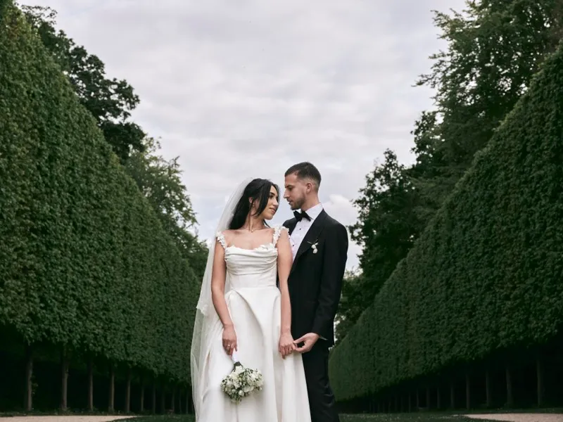 A bride and groom stand close on a lawn between towering clipped hedges under a grey sky.