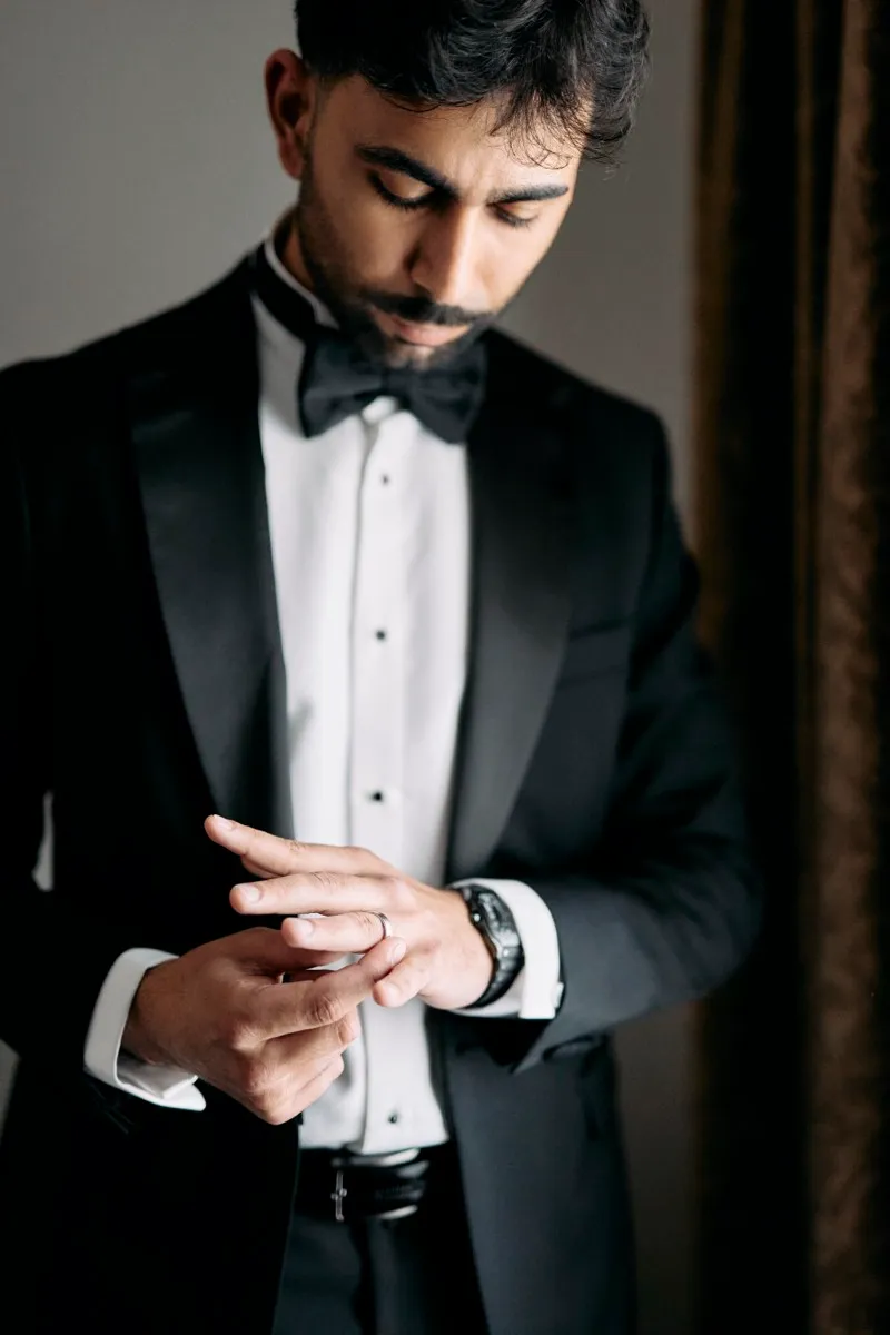 A groom in a black tuxedo and bow tie fastens his cufflink beside a window with dark curtains.