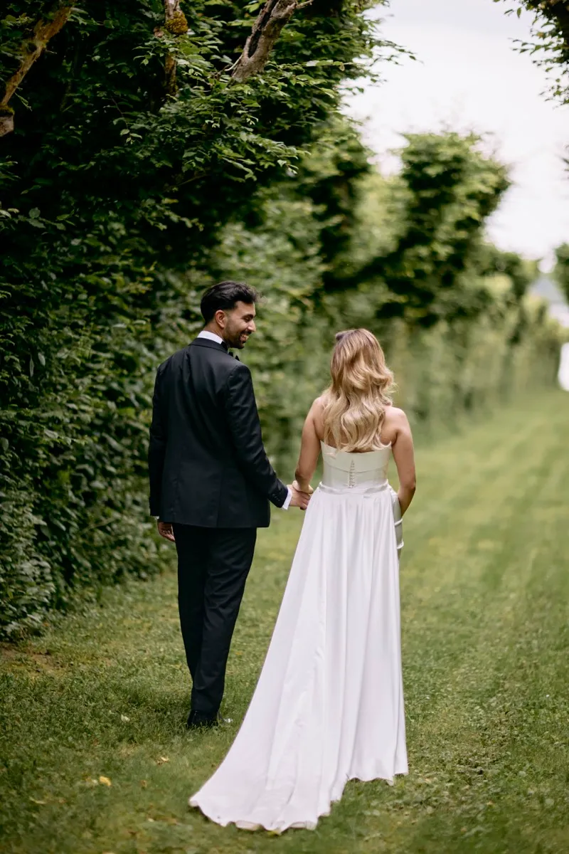 A bride and groom walk away from the camera hand in hand along a hedge-lined lawn, her train trailing on the grass.