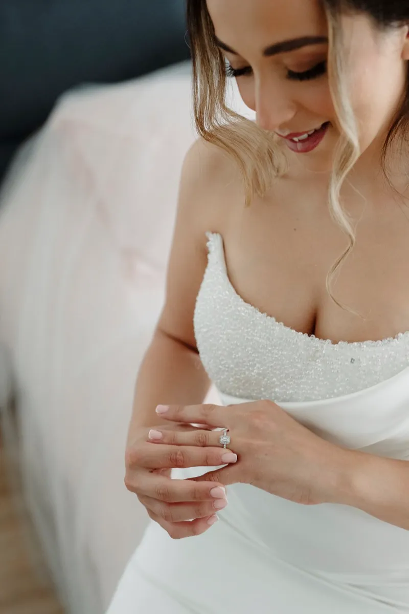 A bride in a beaded strapless gown looks down at the ring on her hand, her veil soft in the foreground.