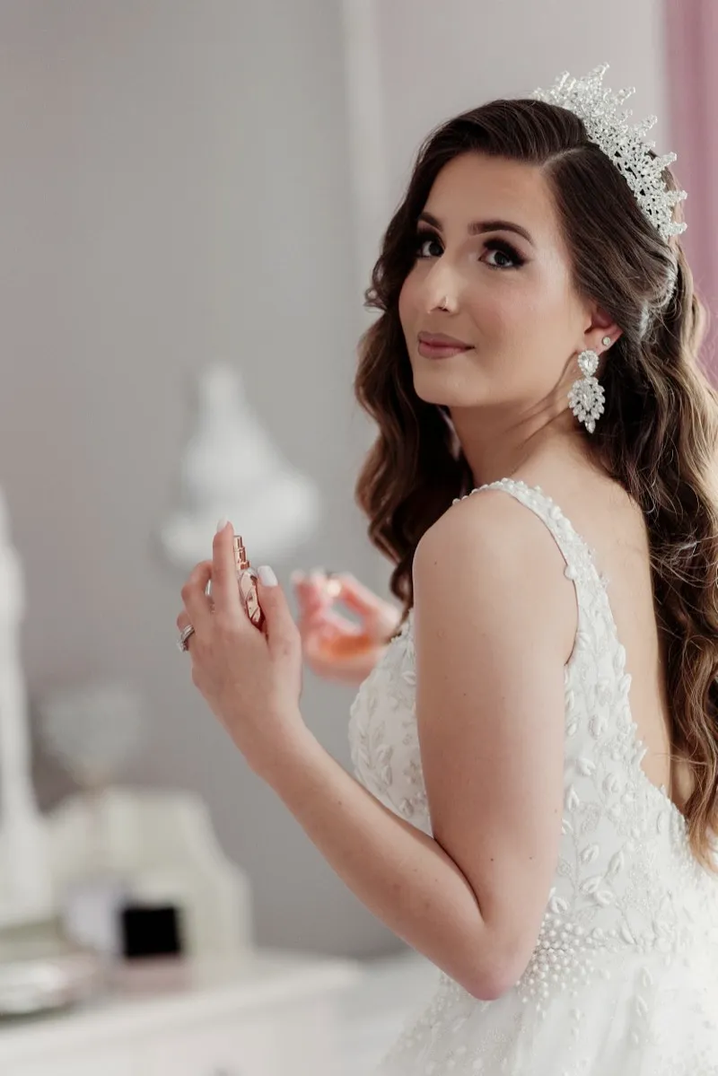 A bride in a beaded gown and crystal headpiece looks over her shoulder while spraying perfume at her dressing table.
