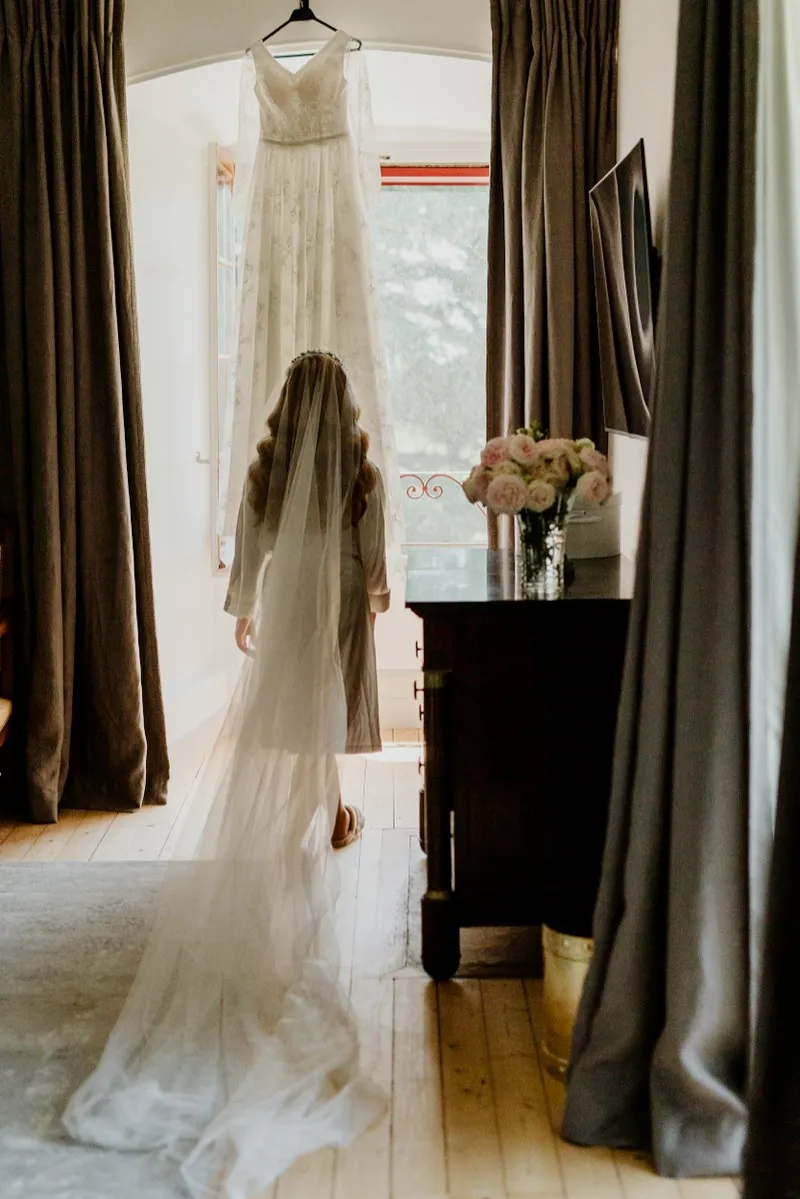A bride stands in her veil at a sunlit window with her gown hanging above her and roses on the dresser.