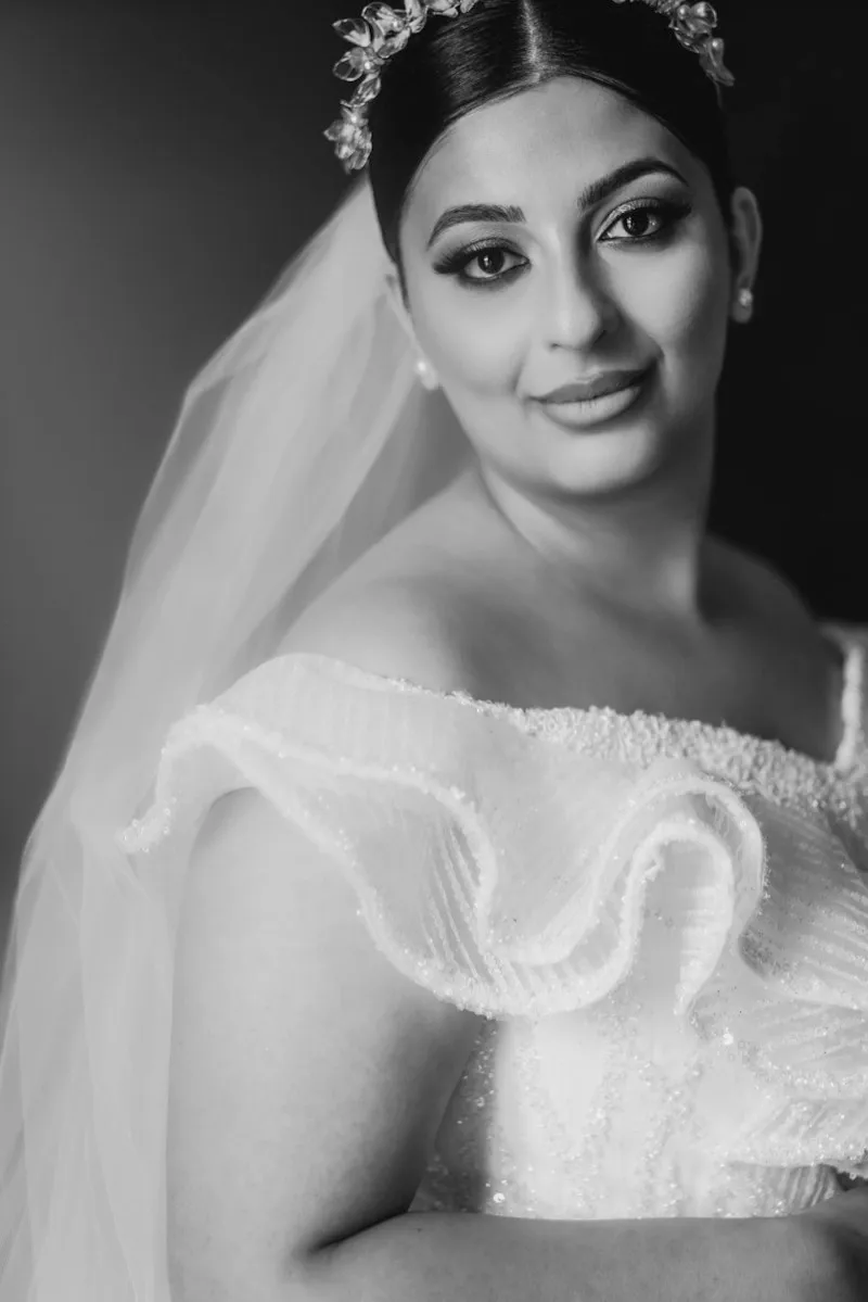 Black and white portrait of a bride in a ruffled off-the-shoulder gown and jewelled headpiece, looking at the camera through her veil.