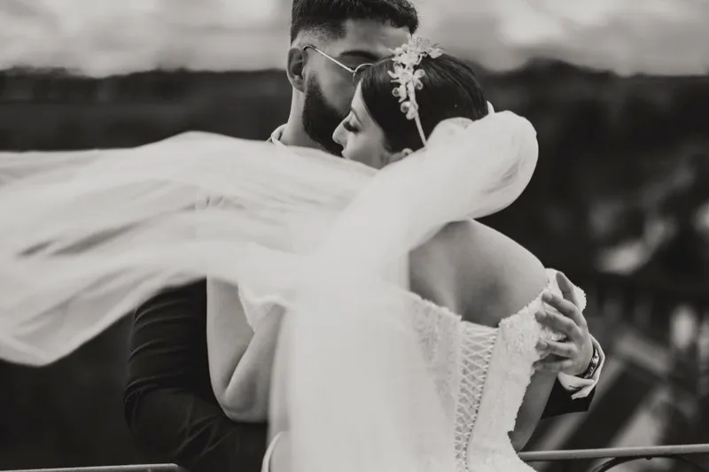 Black and white photograph of a bride's veil blowing across the frame as she embraces her groom on a terrace.