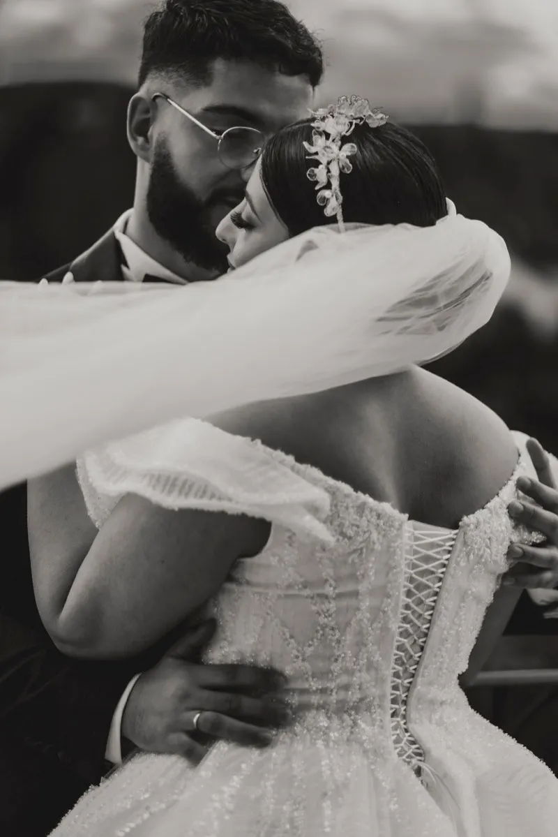 Black and white photograph of a groom in round glasses holding his bride close, her veil wrapped around them both.
