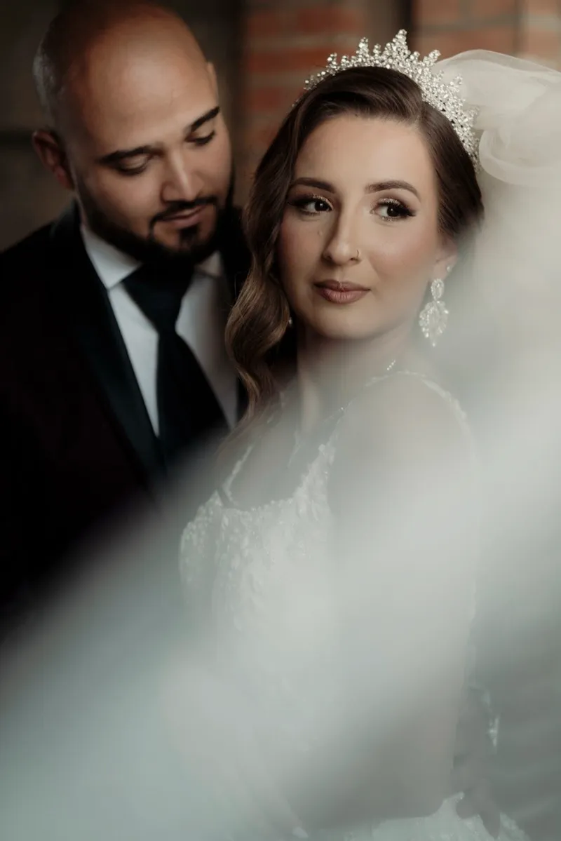 A groom in a dark suit looks down at his bride, who wears a crystal crown and looks toward the camera, seen through her veil.