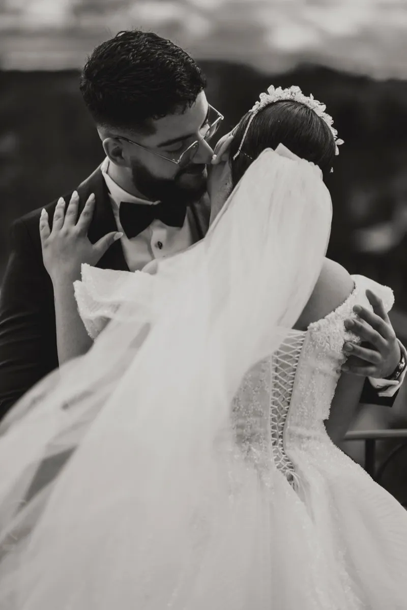 Black and white photograph of a groom in a tuxedo kissing his bride's cheek, her veil lifted by the wind.