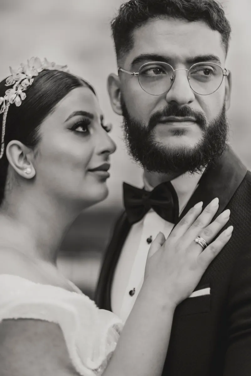 Black and white portrait of a bride with a beaded headpiece resting her hand on the chest of her groom, who wears round glasses and a bow tie.
