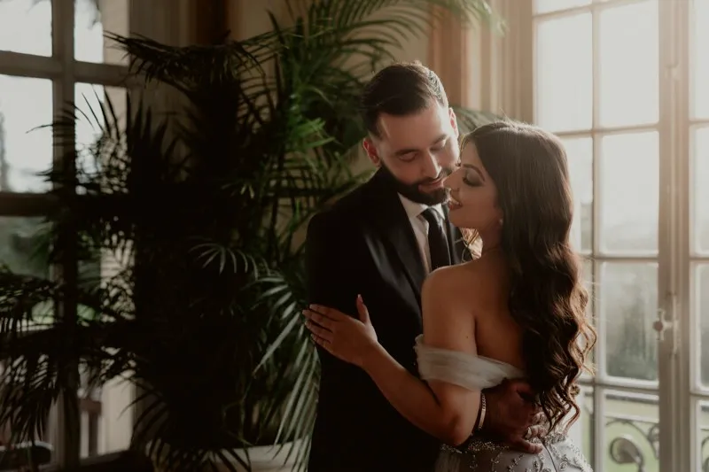 A groom kisses his bride beside a tall window with palms behind them, her off-the-shoulder gown catching the light.