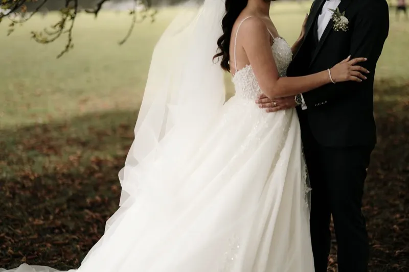 A bride in a tulle gown and long veil holds her groom's lapel on a lawn scattered with autumn leaves.