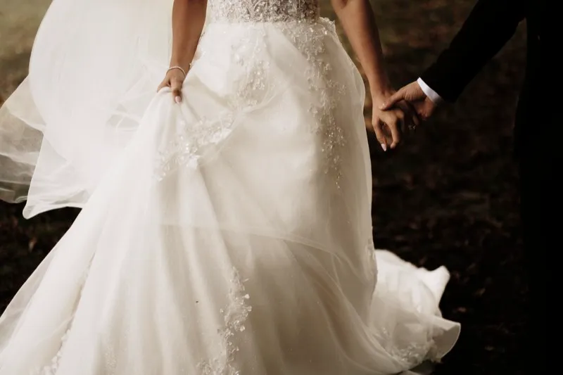 Detail of a bride lifting her tulle skirt as she walks hand in hand with her groom across a lawn.