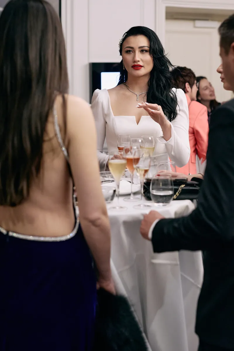 A bride in a long white backless dress, seen from behind, chats with guests at a cocktail table.