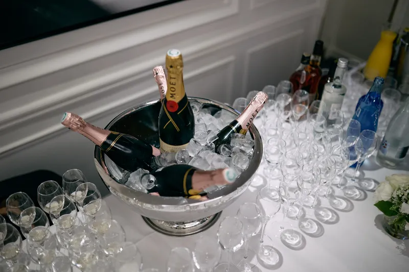 A bottle of champagne resting in a silver bucket of ice beside rows of empty glasses.