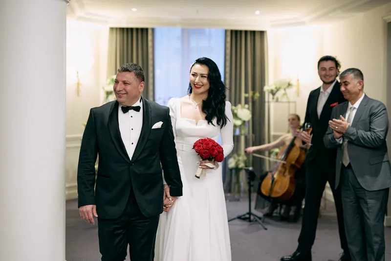 A bride holding red roses and a groom in a black tuxedo enter the reception room as a string trio plays behind them.