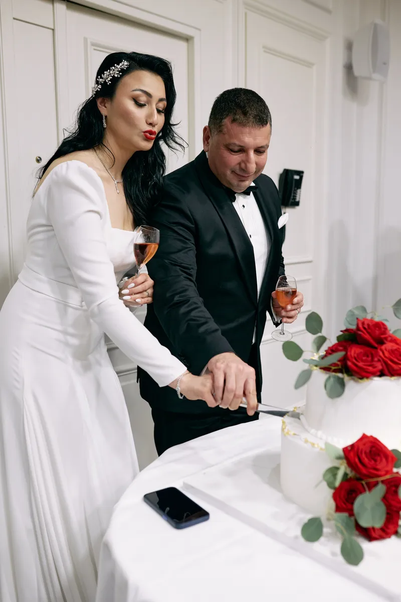 The bride and groom cut their wedding cake together, red roses beside it.